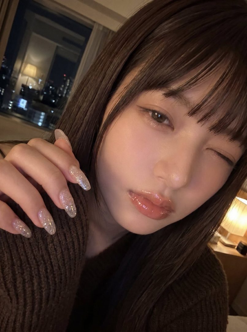 A close-up selfie of a woman with long brown hair and bangs, wearing a brown sweater, winking at the camera.