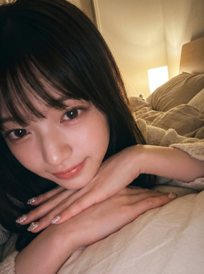 A close-up selfie of a young woman with long black hair and bangs, resting her chin on her hands in a cozy bedroom setting.