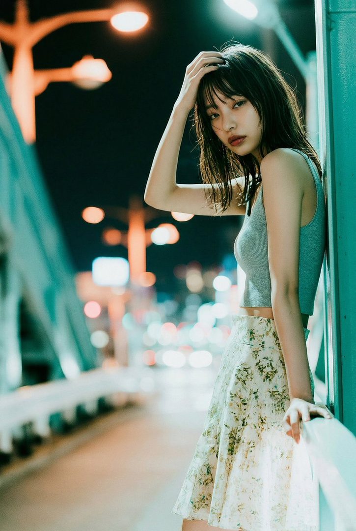 A young woman with dark hair and bangs poses on a bridge at night, with city lights blurred in the background.