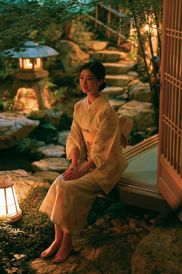 A young woman in a kimono sits barefoot in a Japanese garden at night, illuminated by soft lanterns.