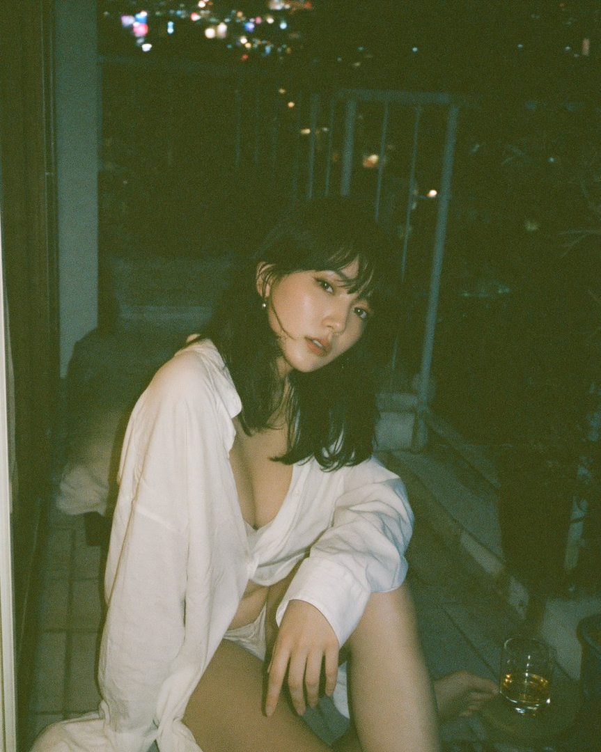 A woman with long black hair and bangs sits indoors at night, wearing an unbuttoned white blouse, with city lights visible in the background.