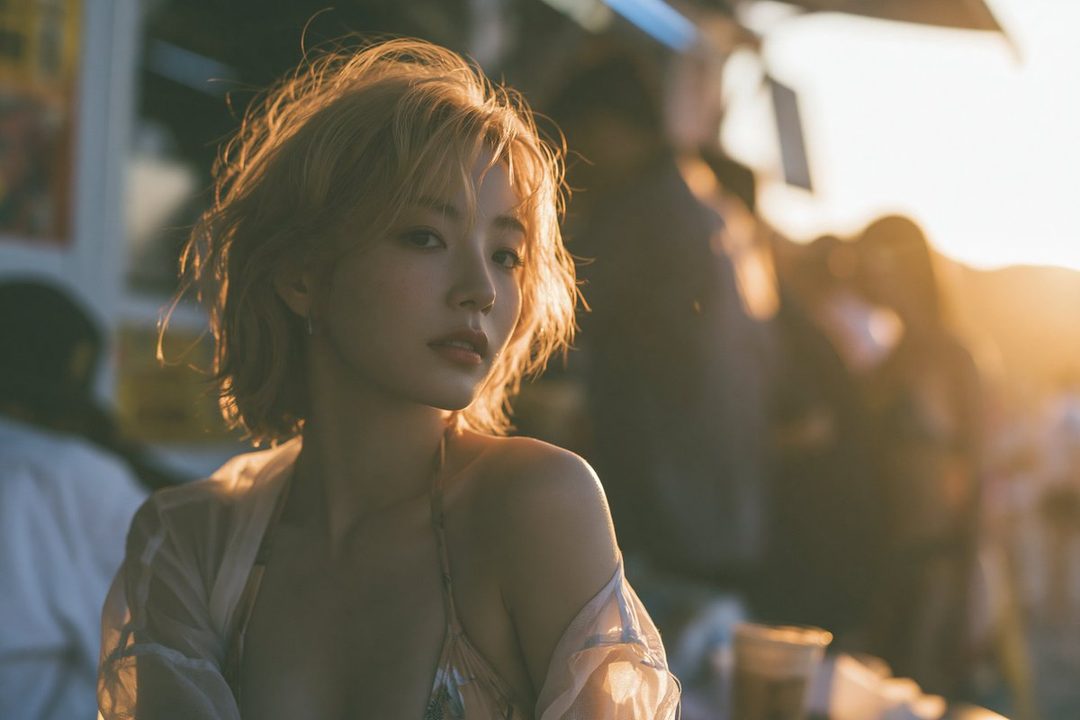 A woman with short blonde hair and a light top over swimwear looks thoughtfully into the distance during golden hour.