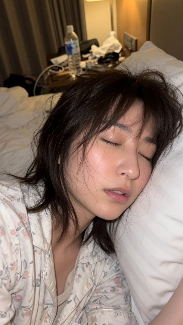 A woman with brown hair and bangs is sleeping peacefully on a white pillow in a bedroom, wearing floral pajamas.