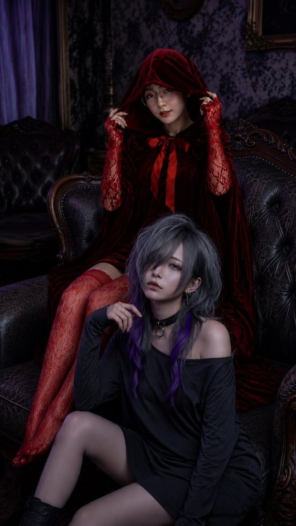 Two women posing in a dark, gothic-style indoor setting, one in a red hooded cape and the other with silver and purple hair in a black dress.