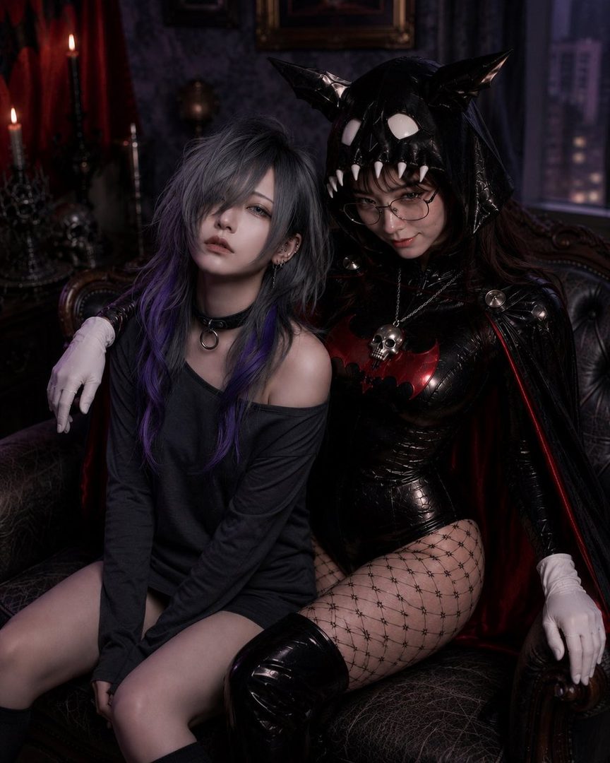Two women in gothic-inspired cosplay, one with silver and purple hair in a black off-shoulder top, and the other in a black latex bat-themed costume with fishnet stockings, sitting on a dark leather couch.