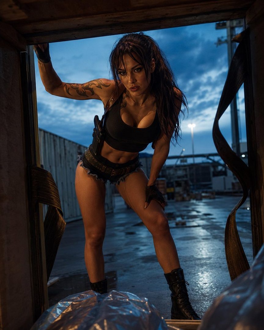 A woman in a black crop top and denim shorts, resembling Lara Croft, poses in a dark, industrial outdoor setting at night.