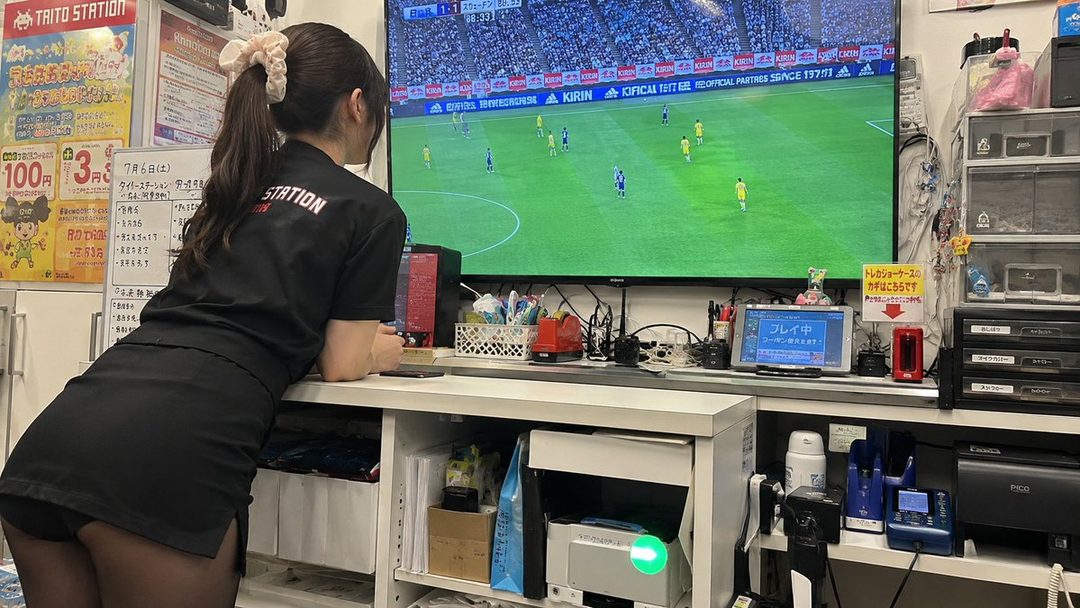 A woman in a black uniform with a ponytail watches a soccer game on a large screen in an indoor setting.