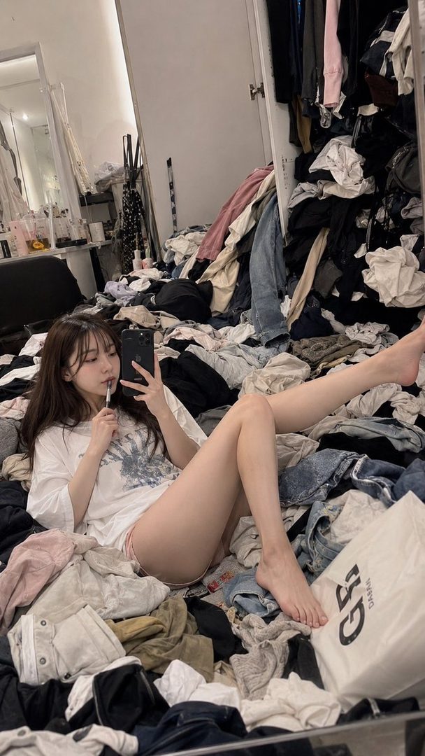 A young woman in a white t-shirt and shorts lies on a pile of clothes, taking a selfie with her phone.