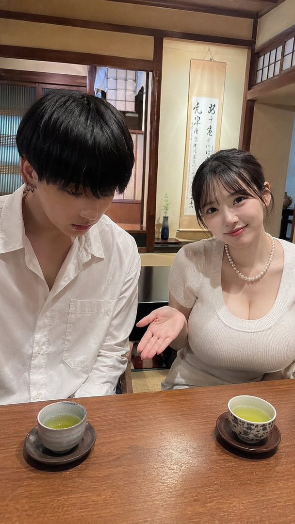 A young woman with a pearl necklace and a man are sitting at a wooden table with two cups of tea in a traditional Japanese setting.