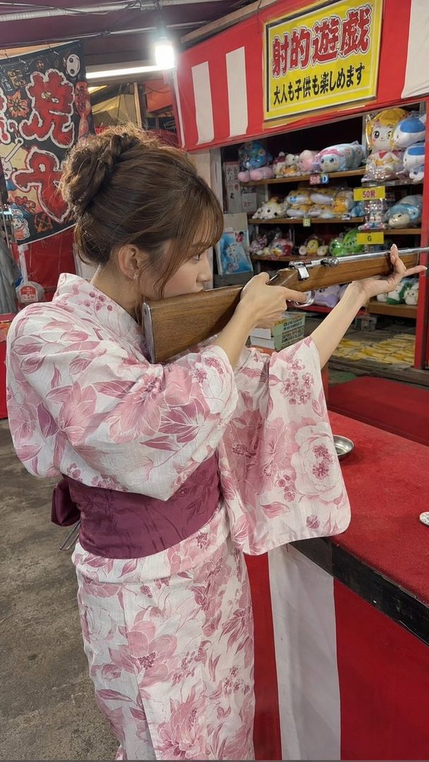 A woman in a pink floral yukata aims a rifle at a shooting game booth at a festival.
