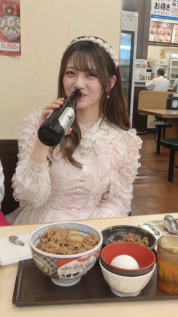 A young woman in a pink frilly dress drinks from a bottle while sitting at a table with a bowl of gyudon.
