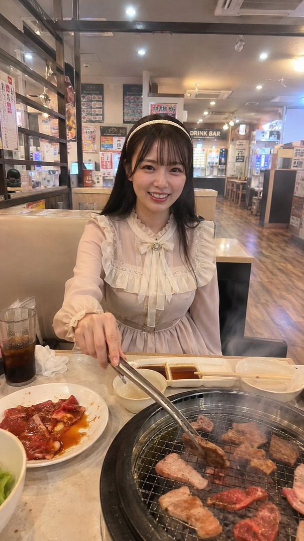 A smiling woman with long black hair and bangs, wearing a light-colored ruffled dress, grilling meat at a restaurant.