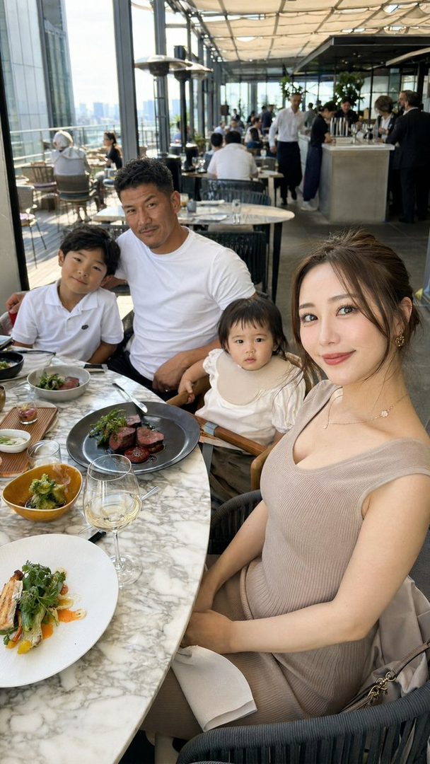A family of four, including a woman in a beige dress, a man, and two children, are seated at a table in a restaurant with a city view.