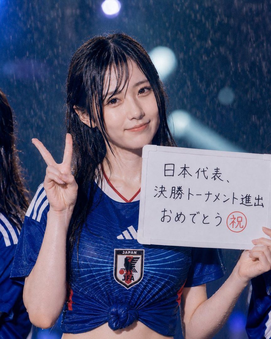 A young woman with wet black hair, wearing a blue Japan national football team jersey, holds a whiteboard with Japanese text and makes a peace sign.