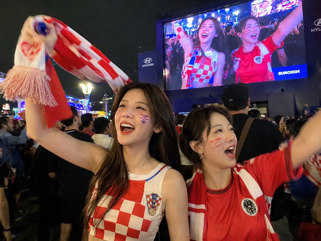 Two young women in sports jerseys cheer excitedly at a night event, with a large screen showing a game in the background.