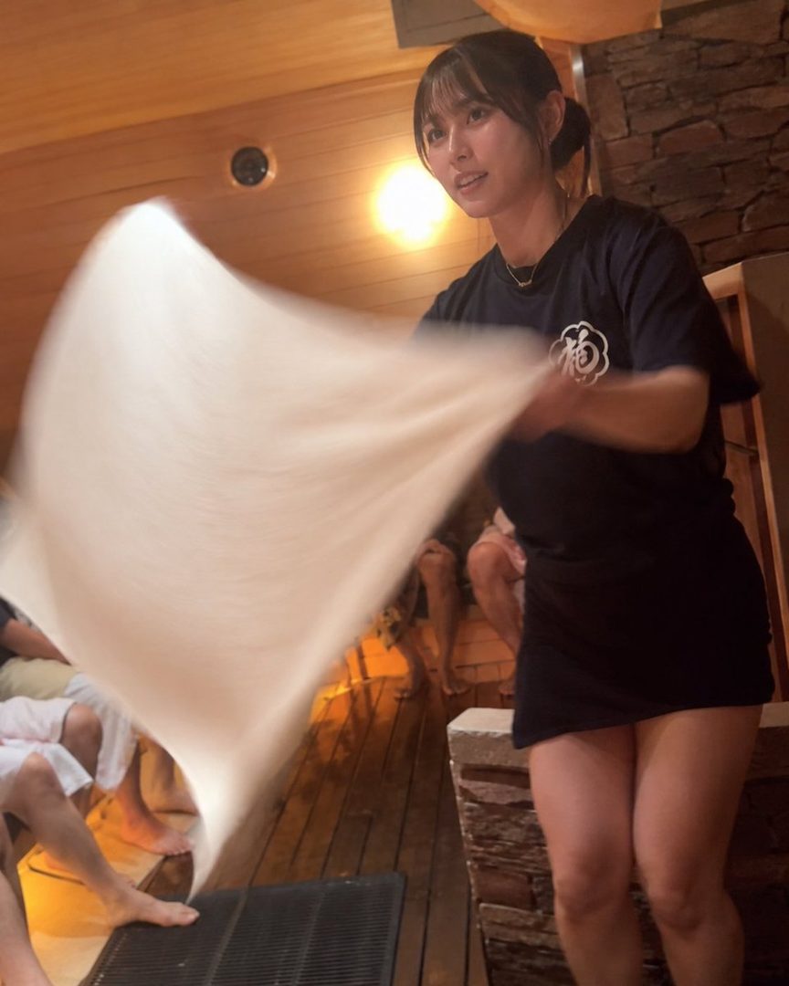 A young woman in a dark t-shirt and skirt is fanning a white cloth in a dimly lit wooden room, possibly a sauna.