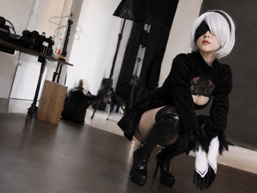 A woman cosplaying as 2B from Nier: Automata, wearing a black dress and blindfold, crouching in a studio setting.