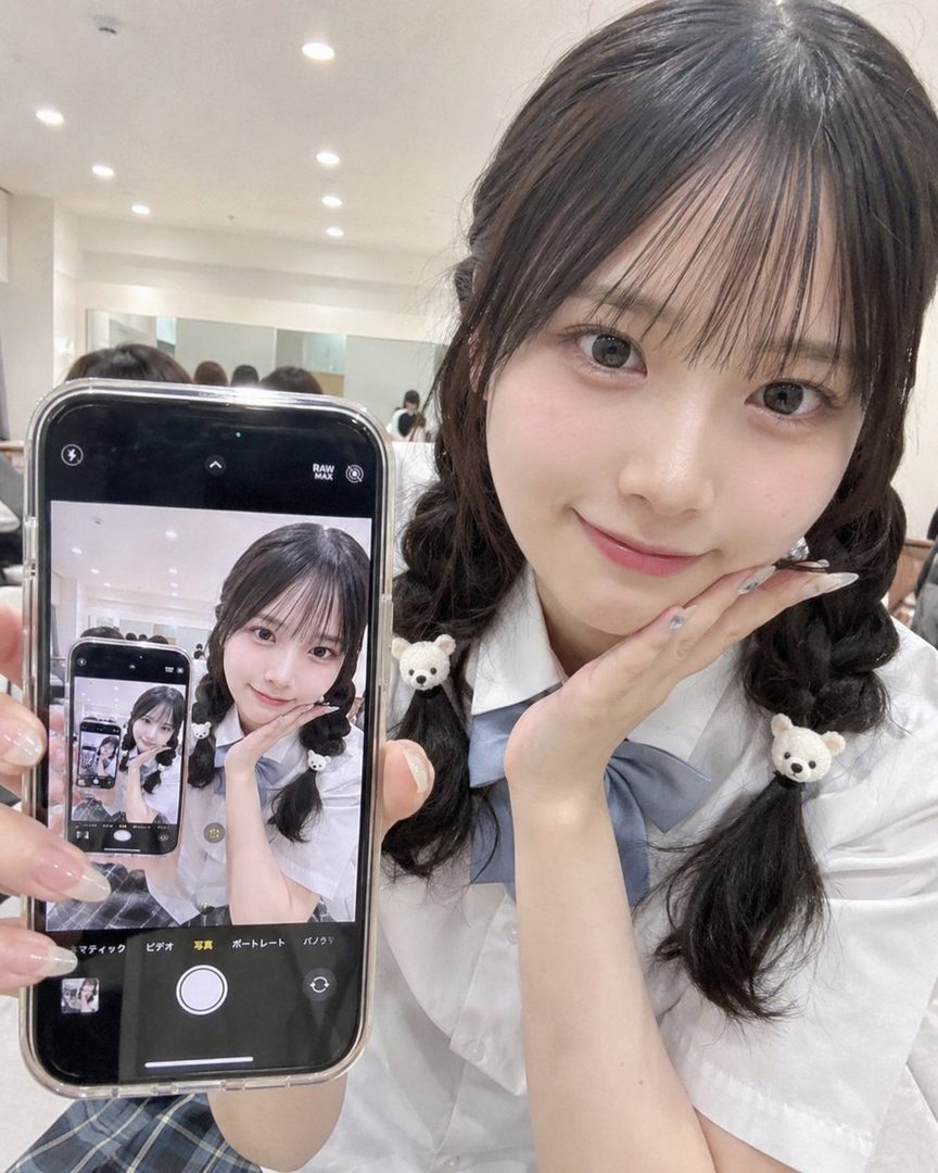 A cute East Asian girl in a school uniform takes a selfie, holding up her phone to show a previous selfie of herself.