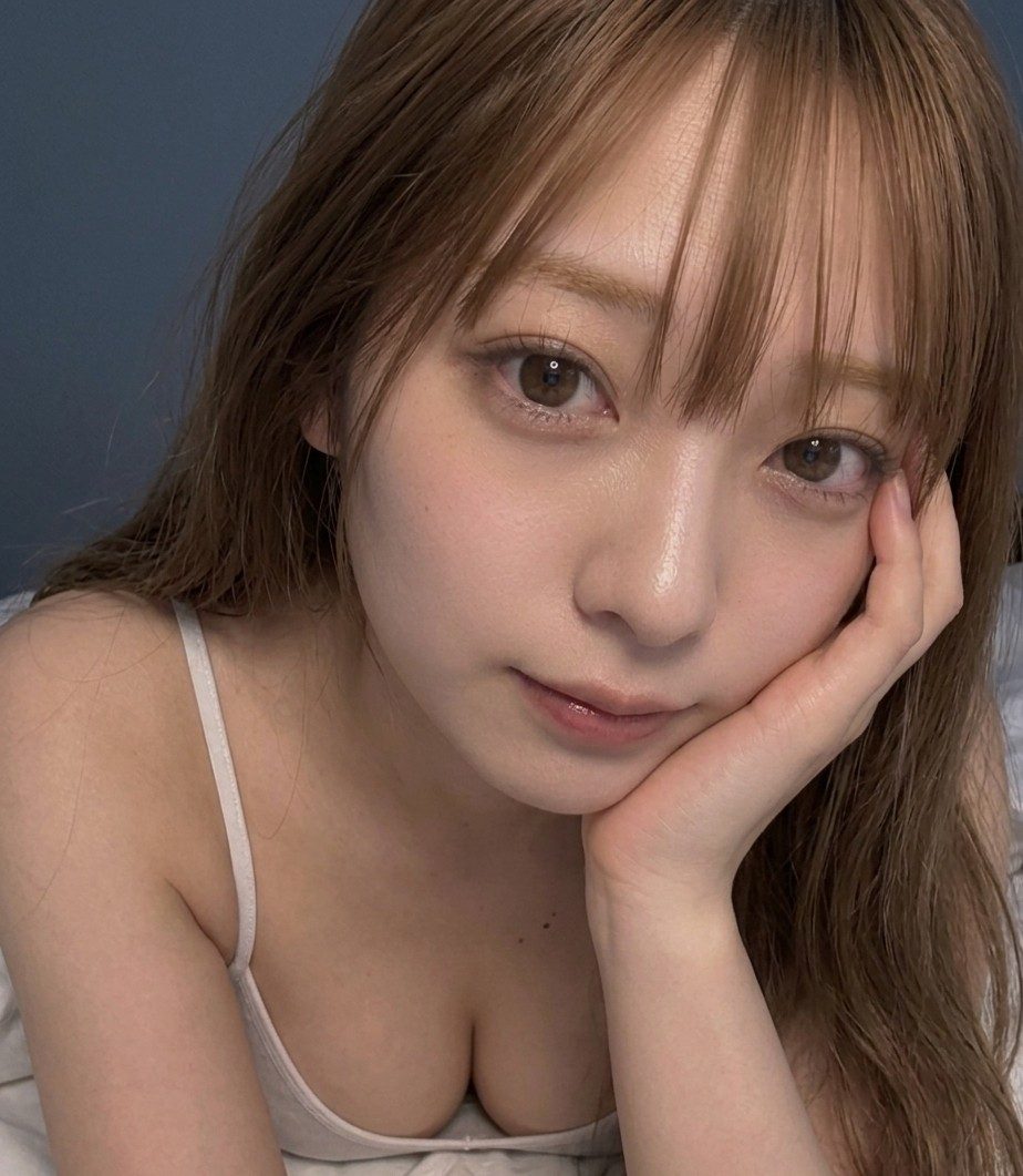 A close-up portrait of a young woman with brown hair and bangs, wearing a white camisole, resting her chin on her hand.