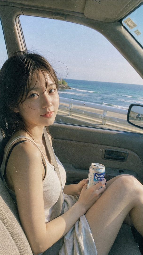 A young woman with brown hair sits in a car, holding a can, with the ocean visible outside the window.