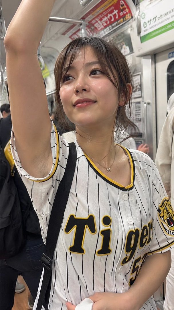A young woman with brown hair and bangs, wearing a white and black striped baseball jersey, stands in a train.