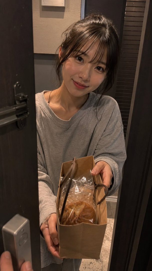 A young woman with brown hair and bangs, wearing a grey long-sleeved shirt, smiles while holding a paper bag with bread inside, peeking from behind a door.