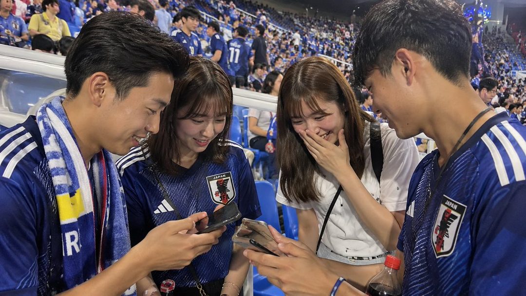 Four young people in blue Japan soccer jerseys are looking at smartphones and laughing in a stadium.