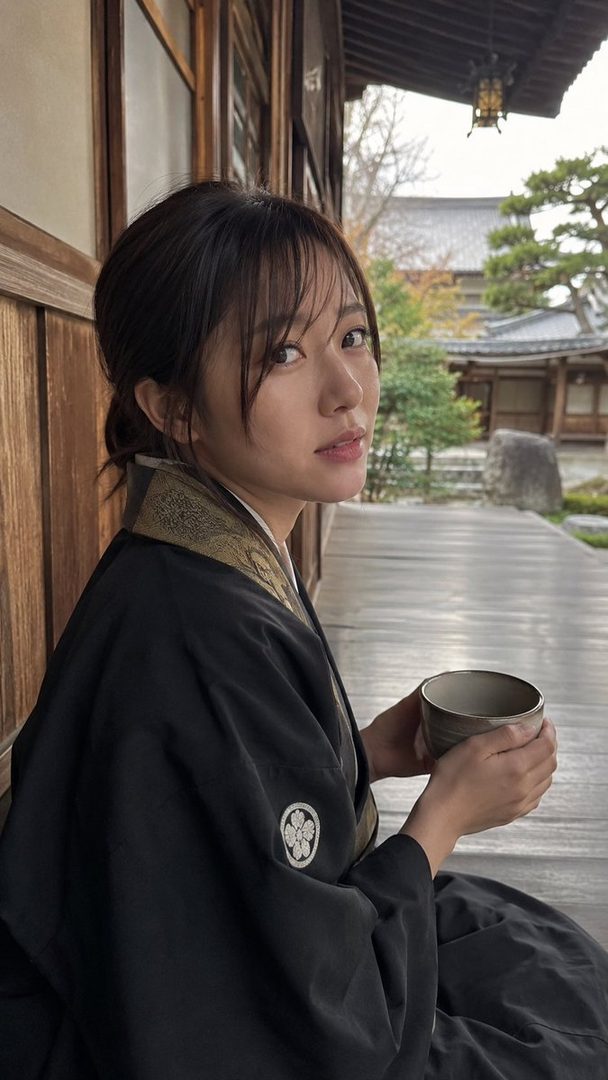 A woman in a black kimono sits on a wooden porch, holding a teacup and looking over her shoulder.