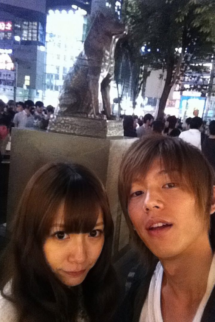 A young Japanese couple takes a selfie in front of the Hachiko statue at Shibuya Crossing at night.