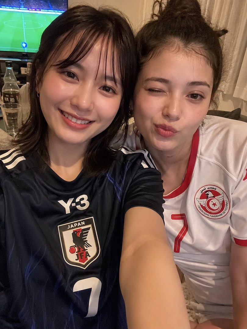 Two young women in soccer jerseys take a selfie indoors, one winking and the other smiling.