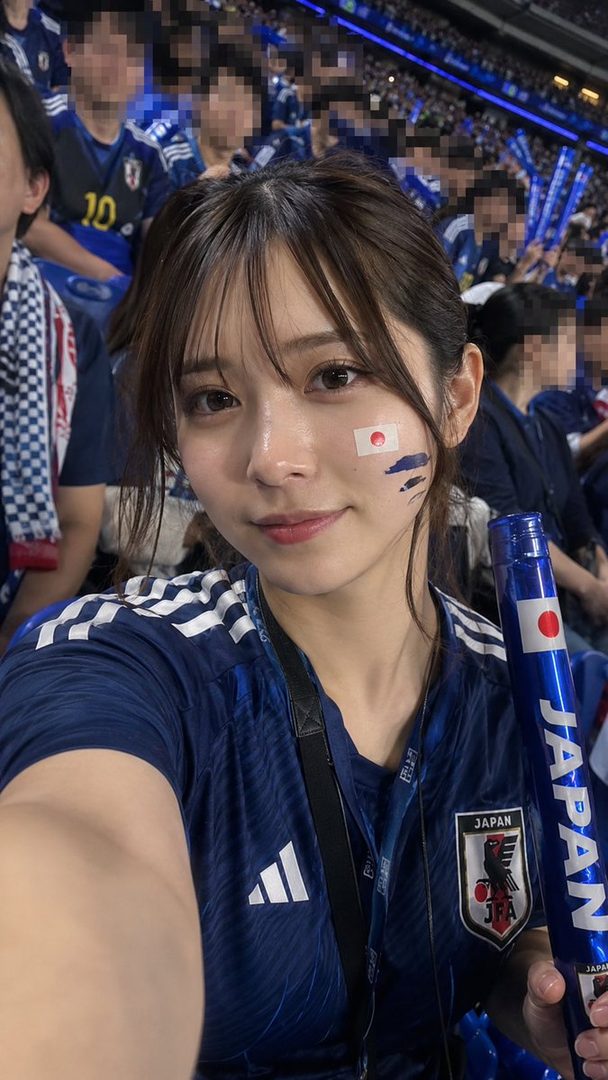A young woman takes a selfie at a soccer match, wearing a Japan national team jersey and face paint.