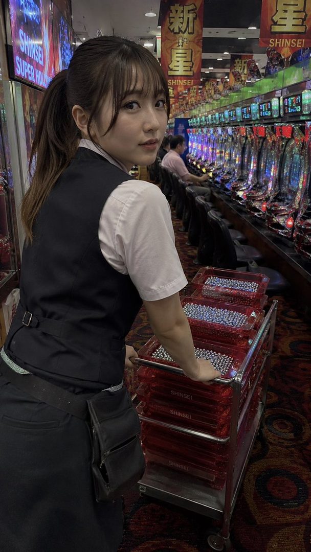 A young woman with brown hair in a ponytail and bangs, wearing a white blouse and black vest, looks over her shoulder while pushing a cart of pachinko balls in a brightly lit pachinko parlor.