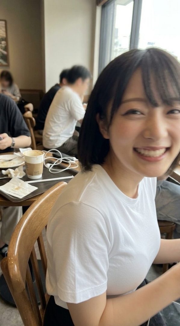 A smiling East Asian woman with short black hair and bangs is sitting in a cafe, wearing a white t-shirt.