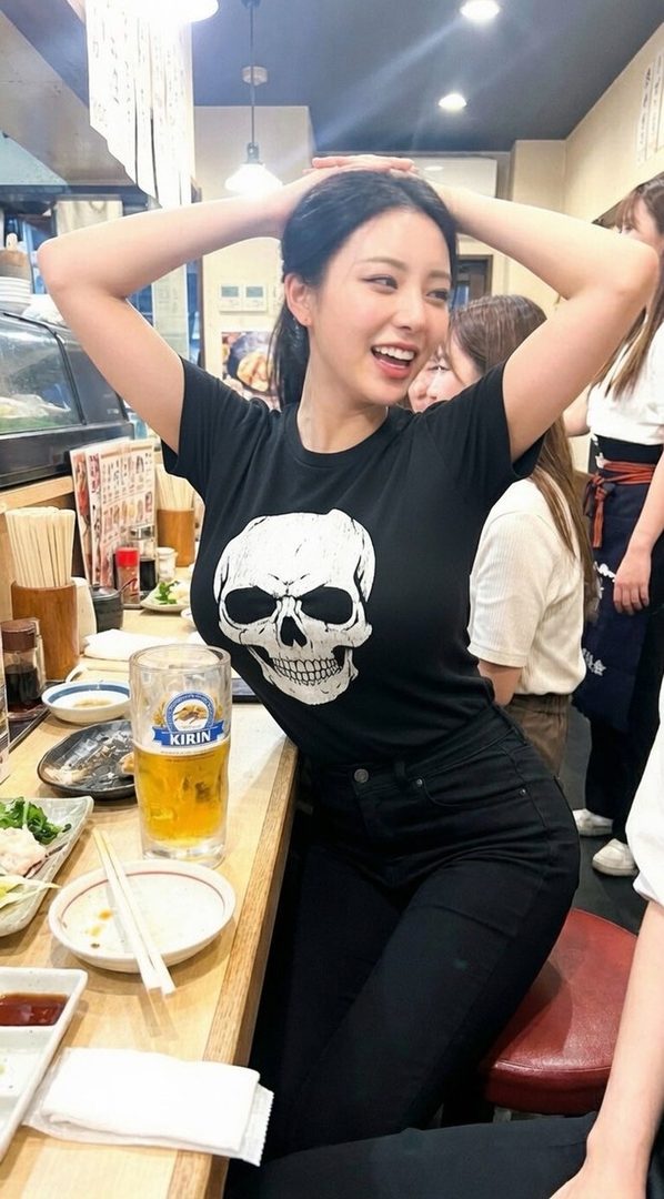 A young woman with black hair wearing a black t-shirt with a skull design, smiling and posing in an izakaya with a glass of beer on the counter.