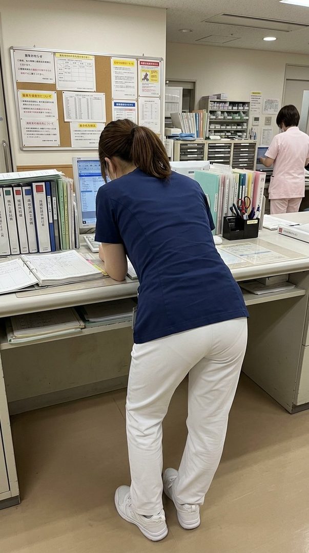 A woman with brown hair tied in a ponytail, wearing a blue top and white pants, leans over a desk in an office setting, looking at a computer screen.
