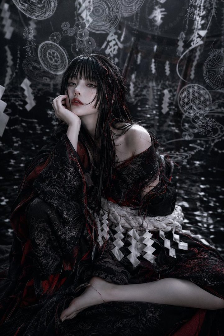 A woman with long black hair and bangs, wearing a dark kimono, sits against a dark background with white patterns.