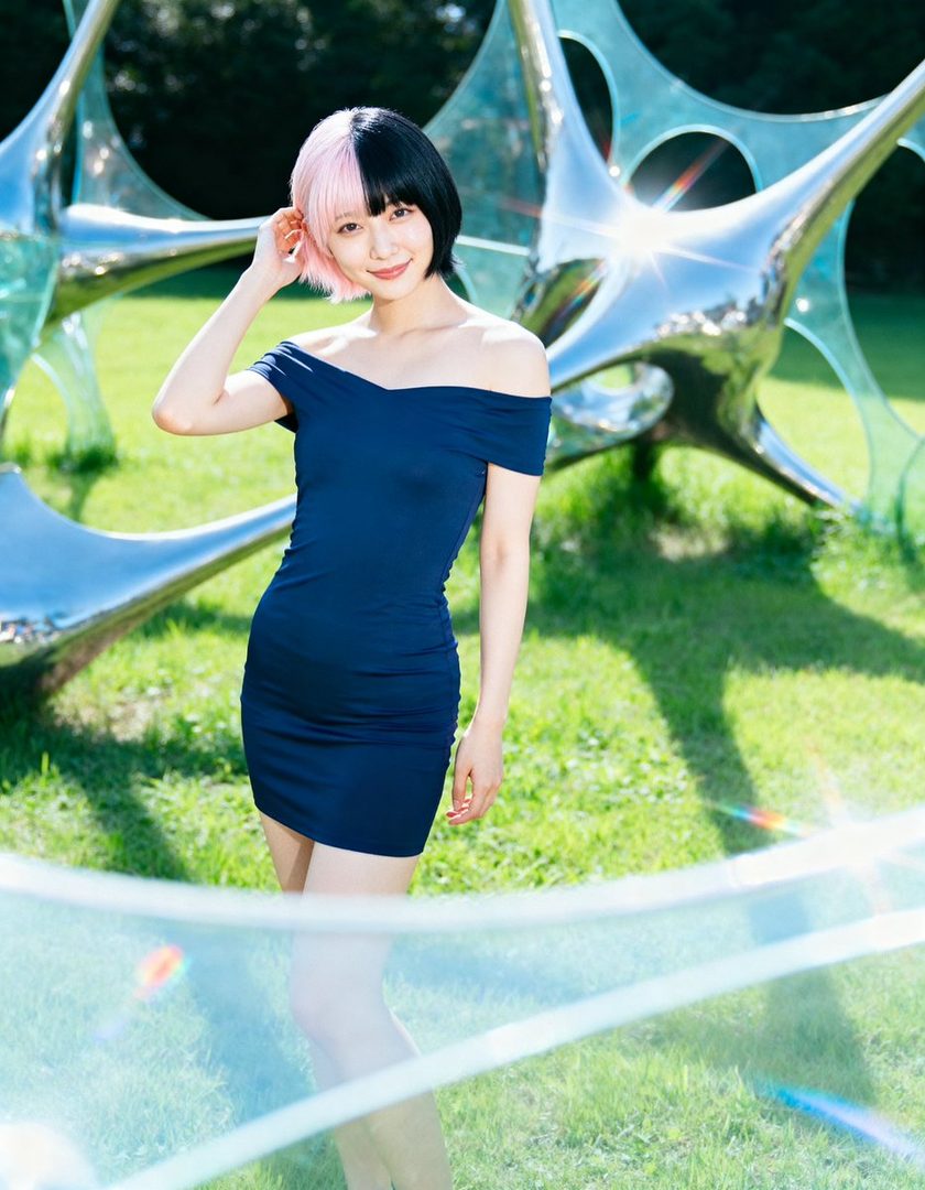 A woman with split pink and black short hair wears a navy off-shoulder dress outdoors, smiling gently.