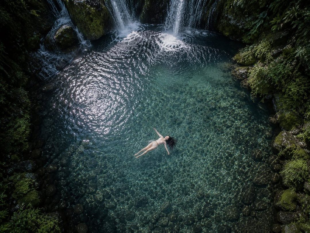 An aerial view of a woman floating on her back in a clear blue natural pool with waterfalls in the background.