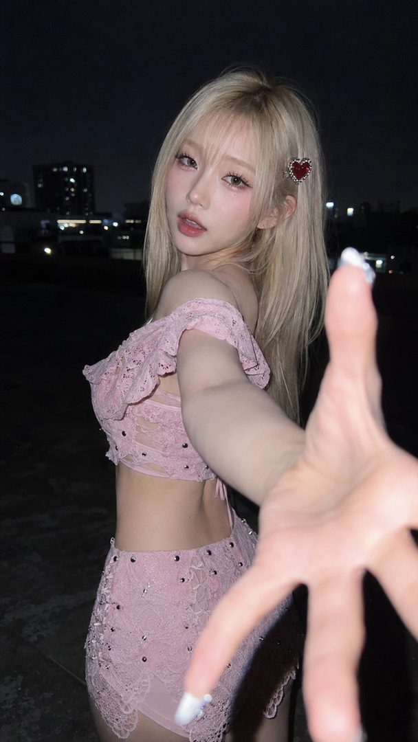 A young woman with blonde hair and a pink lace outfit reaches out towards the camera at night.