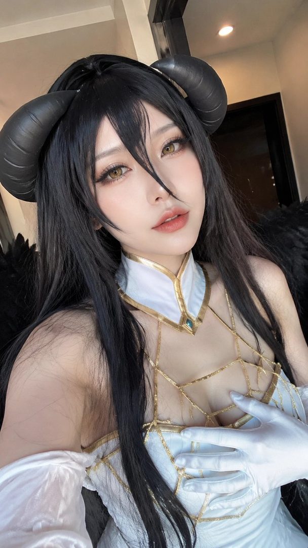 A woman cosplaying as Albedo from Overlord, wearing a white and gold dress with black horns and wings.