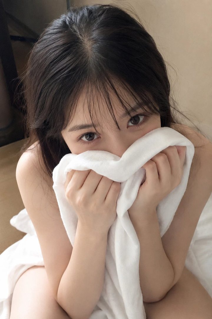 A young woman with dark hair covers her mouth with a white towel, looking directly at the camera.