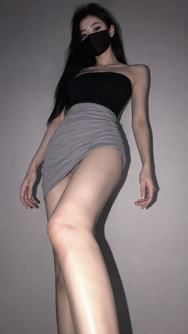 A woman with long black hair and a black face mask wears a black tube top and a gray ruched mini skirt, posing indoors.