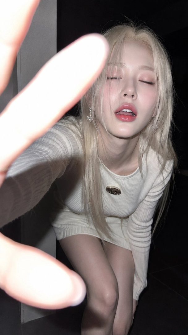 A blonde woman in a white dress takes a close-up selfie with her eyes closed and lips slightly parted, with a blurred hand in the foreground.