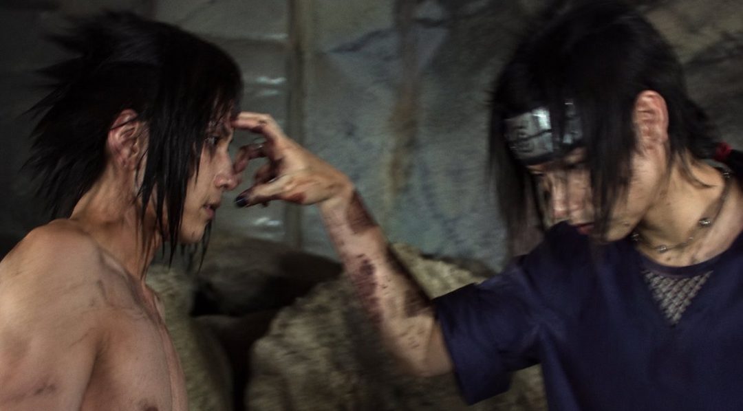 Two men in cosplay, one shirtless and the other wearing a dark top and headband, are depicted in a dramatic indoor scene.