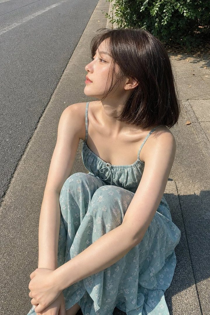 A young woman with short brown hair wearing a light blue floral dress sits on the ground outdoors, looking away from the camera.