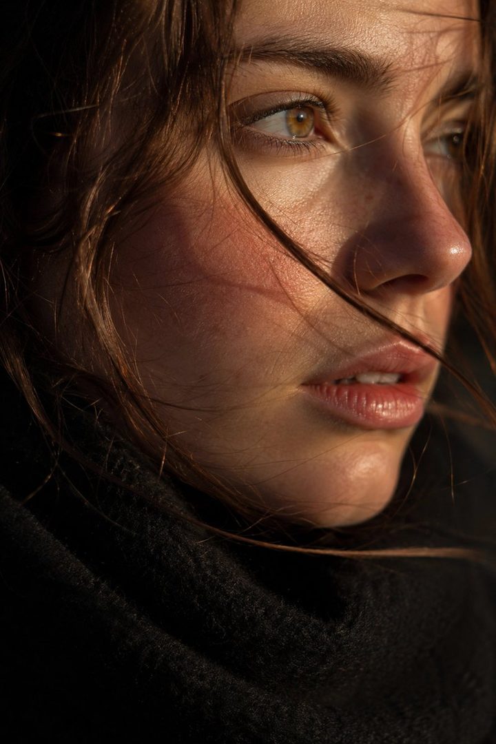 A close-up portrait of a woman with brown hair and warm lighting on her face, wearing a dark sweater.