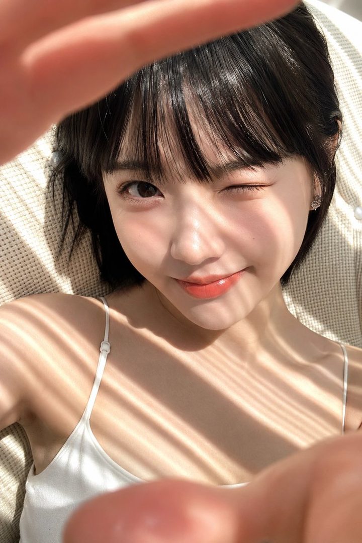 A cute East Asian woman with short black hair and bangs winks at the camera in a sunlit selfie.