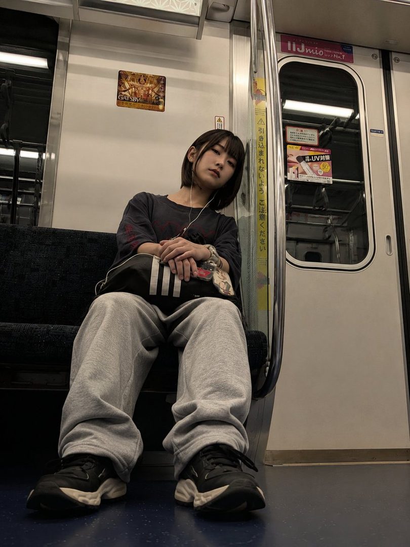 A young woman with short black hair and bangs sits on a train, wearing a dark t-shirt and grey sweatpants, looking towards the camera.
