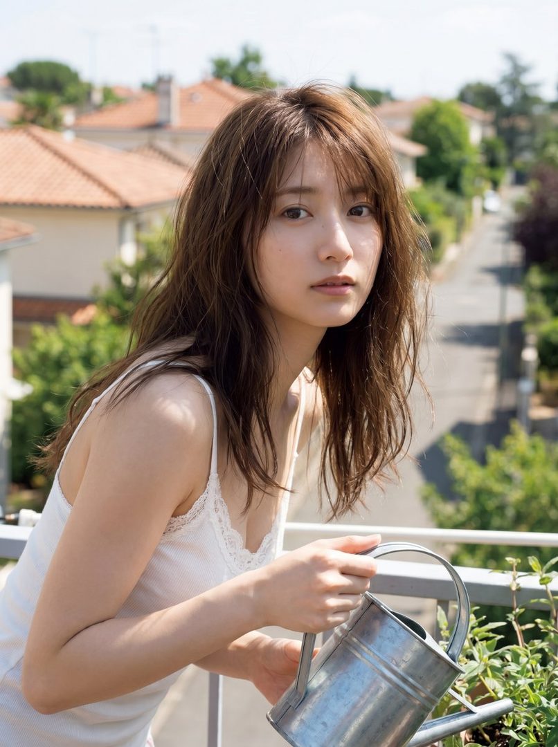 A young woman with long brown hair and bangs, wearing a white camisole, waters plants on a balcony with a metal watering can, overlooking a residential street.