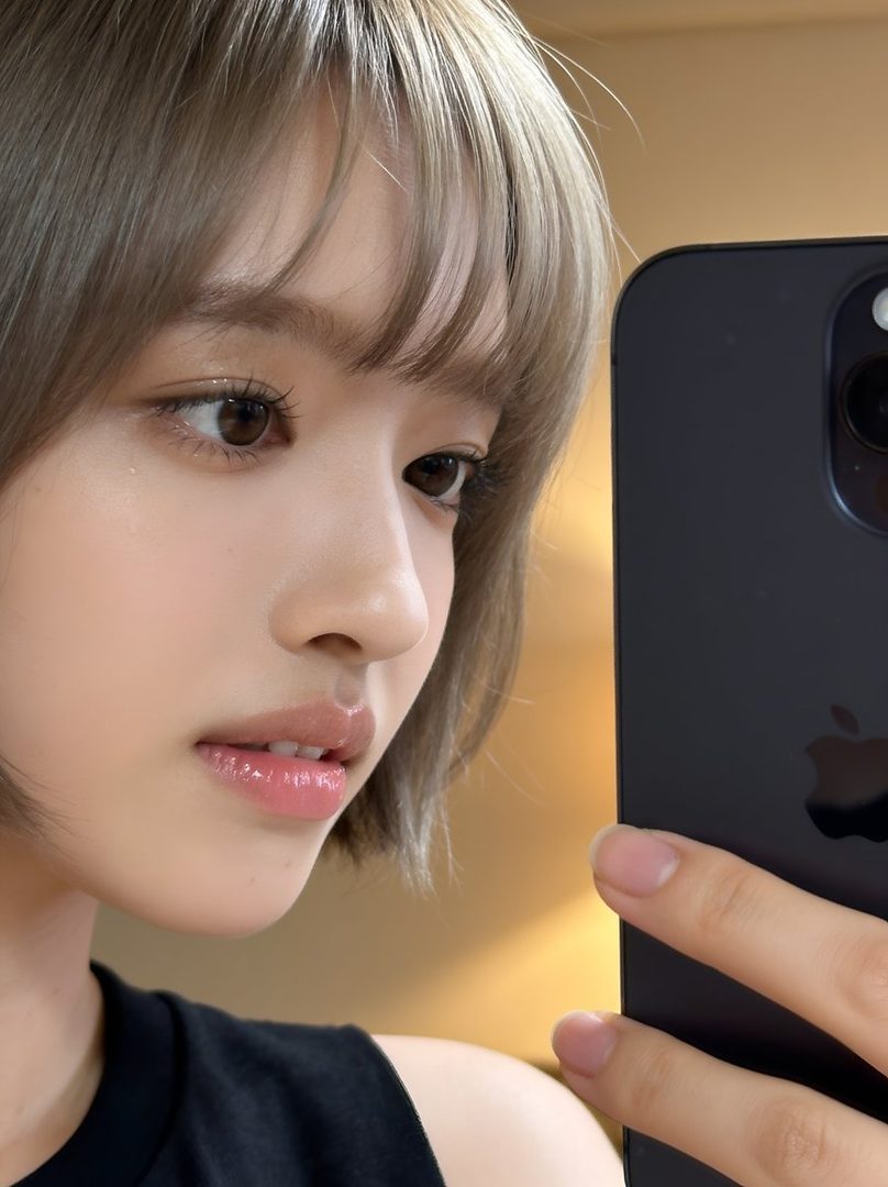 A close-up selfie of a young woman with short blonde hair and bangs, holding a smartphone to take the picture.